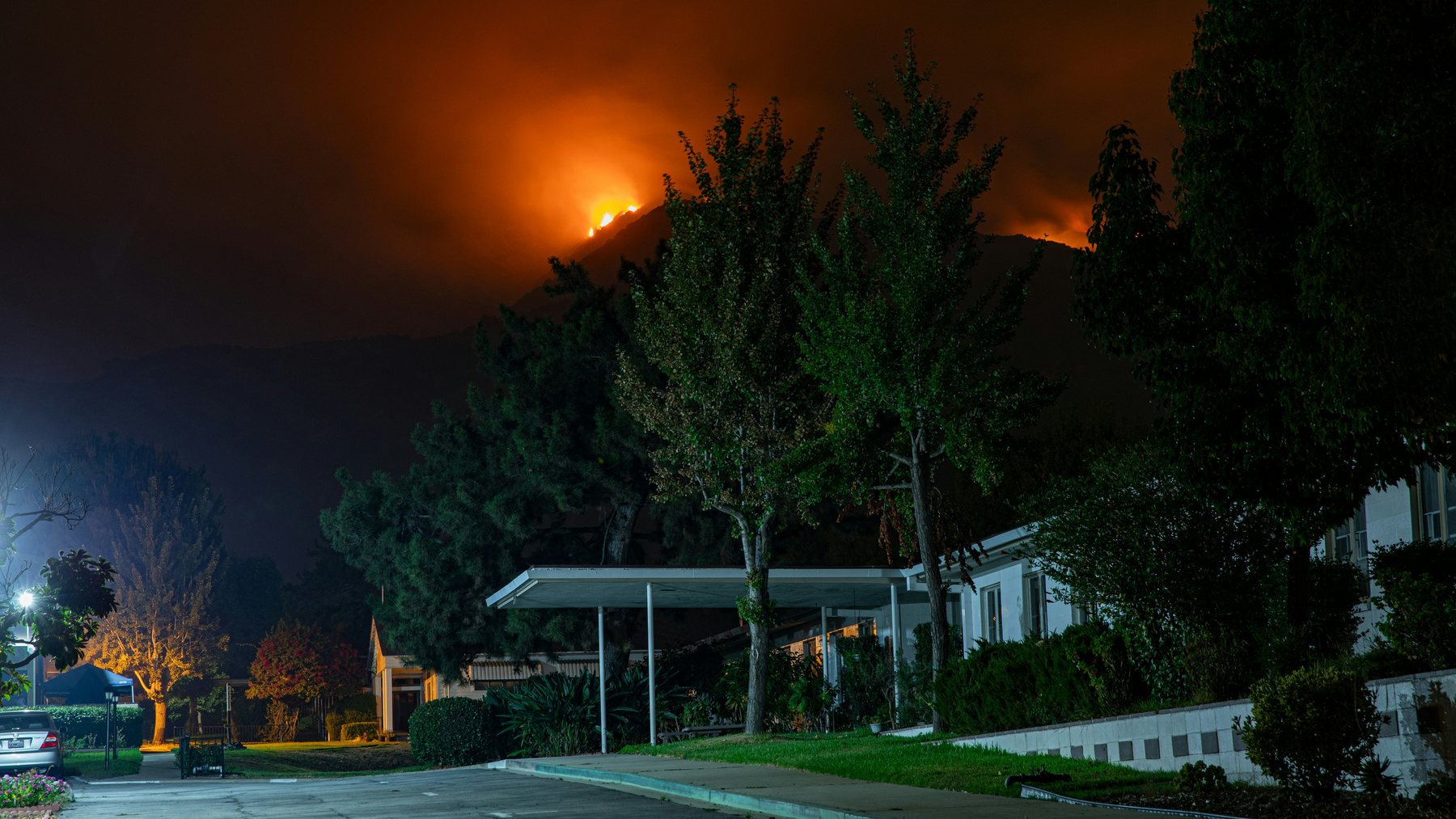 A quiet residential street at night with a wildfire burning on the ridge above the houses