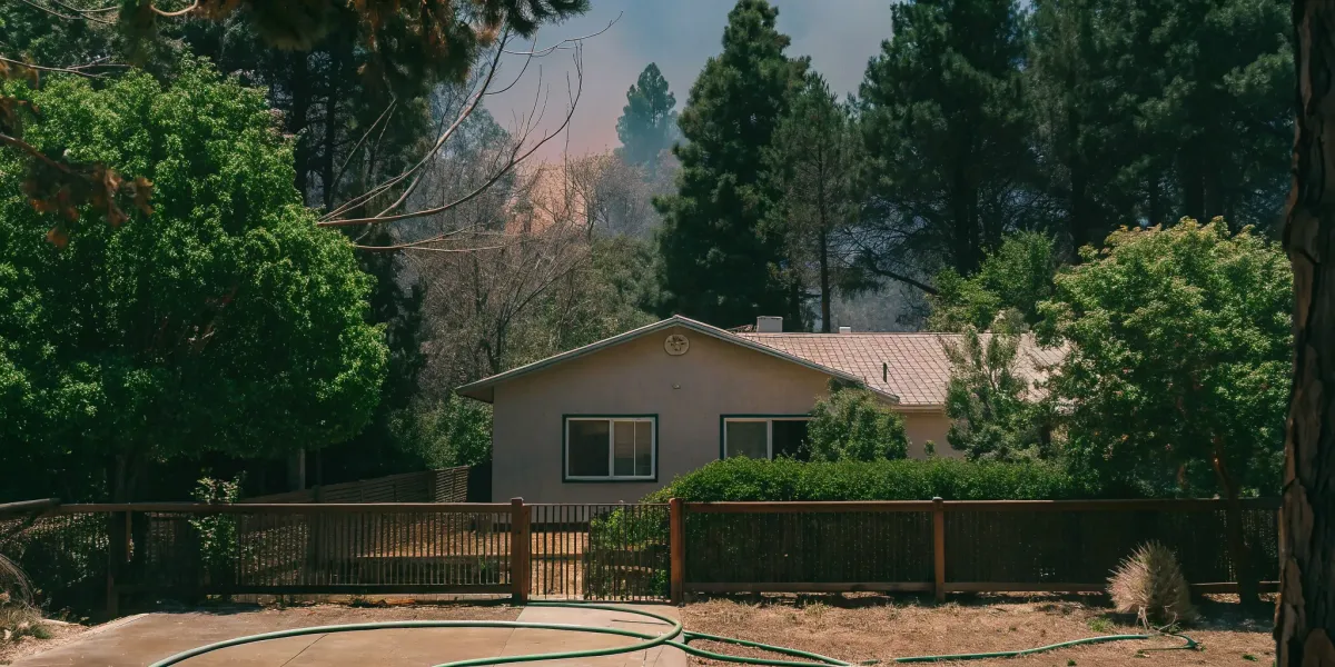 A home's wildfire vulnerability is evident with nearby trees and smoke in the background.