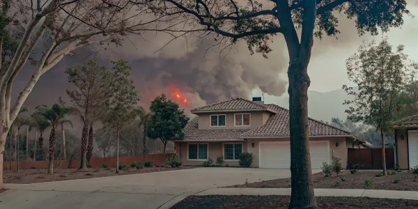 A home in a forested area threatened by an approaching wildfire with smoke and flames.